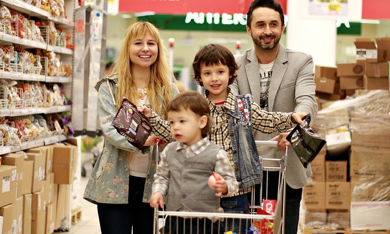 A family with two young children and a shopping cart in a supermarket aisle.