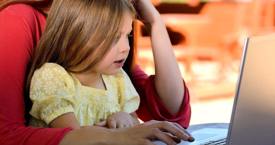 Mother helps her young daughter type on a laptop at a table.