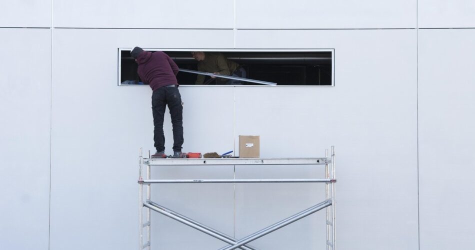Two workers on a mobile scaffold install a panel in an opening on the white facade of a building.